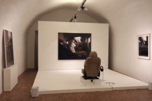 Ausstellungsraum mit Installtion: vor einer Fotografie steht aufgebaut ein Sessel mit Kopfhöhrern daneben 