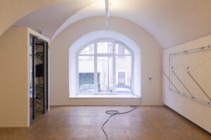 Blick in Ausstellungsraum: Schlauch führt neben einer Wandinstallation,durch ein Fenster, nach draußen 